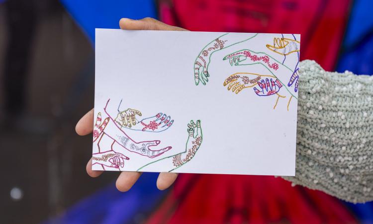 A hand holds a postcard in front of a colourful mural