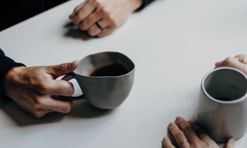 A close up of two hands holding cups of coffee over a white table