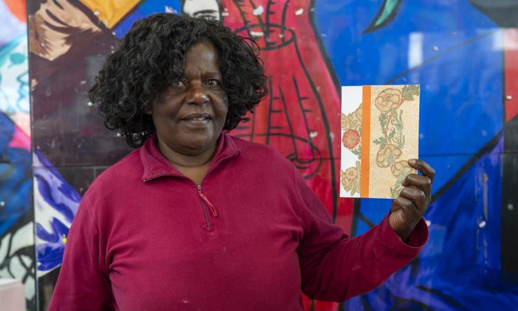 A woman holds a handmade postcard in front of a colourful mural