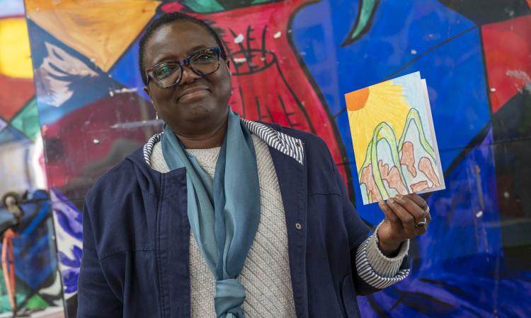 A woman holds a handmade postcard in front of a colourful mural