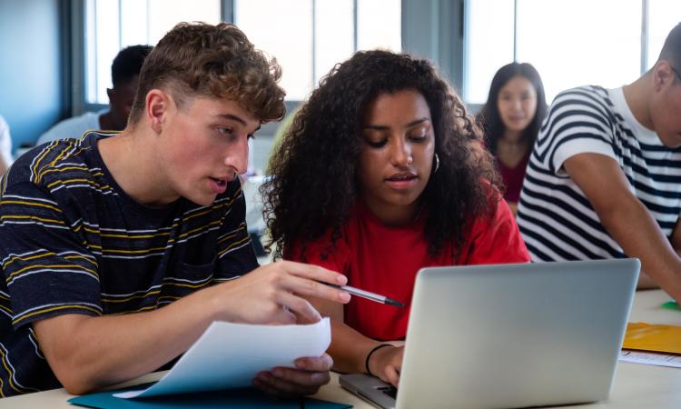 A teenage boy and girl sit at a laptop in a classroom and talk about what's on the screen