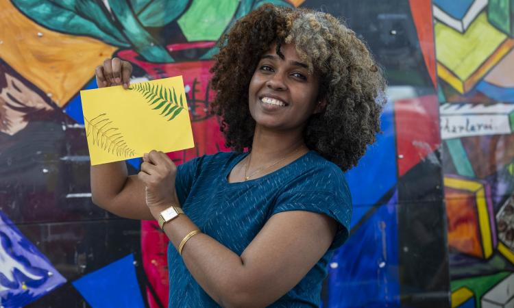 A woman holds a handmade postcard in front of a colourful mural