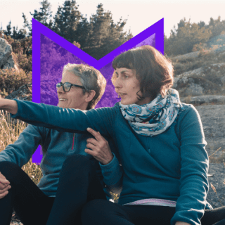 Two women sit in nature, looking to the left of frame where one of them is pointing.